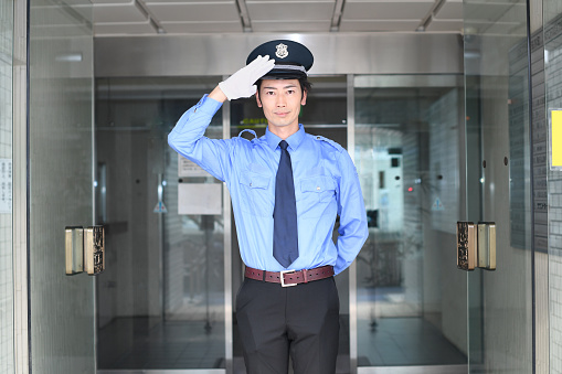 Asian male patrolling security guard wearing hat and uniform saluting in front of building entrance