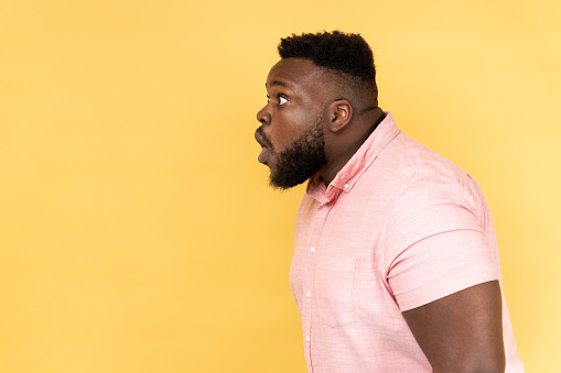 Wow, unbelievable. Side view of man wearing pink shirt has shocked expression, looking at camera with open mouth and amazed big eyes. Indoor studio shot isolated on yellow background.