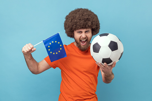 Extremely happy man with Afro hairstyle in T-shirt standing with soccer ball and europe union flag, cheering his favorite team, celebrating victory. Indoor studio shot isolated on blue background.