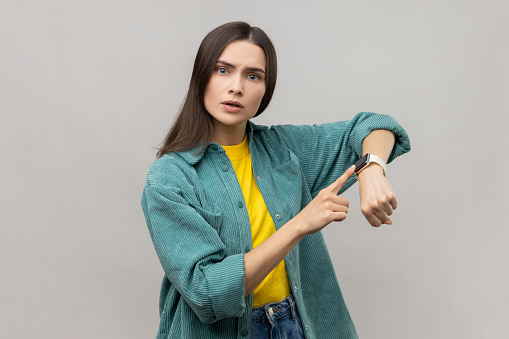 Concerned punctual dark haired woman pointing finger at smartwatch on her wrist, look at time, hurry up and act, wearing casual style jacket. Indoor studio shot isolated on gray background.