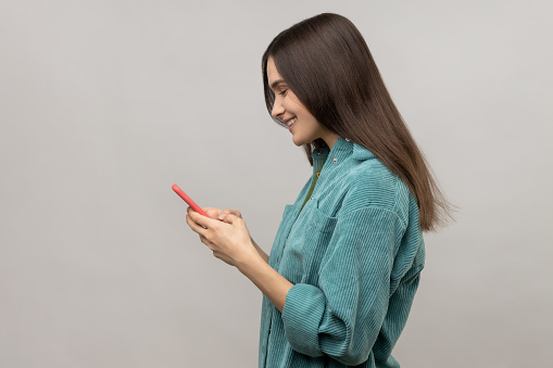 Side view of happy woman reading message on smartphone and smiling, using mobile device for communication, browsing web, wearing casual style jacket. Indoor studio shot isolated on gray background.
