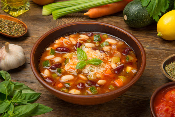 Italian minestrone soup in a brown tarracotta soup bowl on a rustic wooden table with ingredients. stock photo