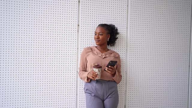 Young black woman using mobile phone and drinking coffee in front of an office building
