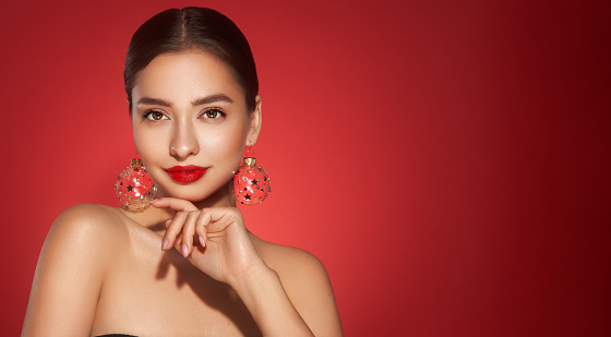 Portrait of beautiful young woman on red background, shows with hands to the side copy space, Christmas toy earrings, shiny glowing skin, Winter holidays concept