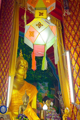 Different seized golden thai buddhas in temple in Chiang Rai province. In foreground are a female one followed by bigger getting ones. In background is big buddha statue with mural behind him. A paper lantern is hanging from ceiling