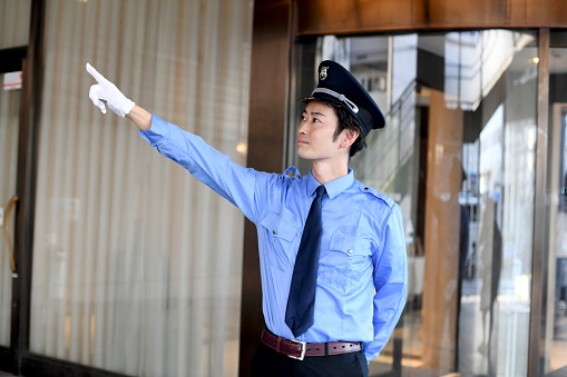 An Asian male patrol security guard wearing a hat and uniform points and checks in front of the building entrance.