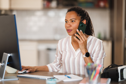 African business woman talking on a smartphone while working on computer from her home office.