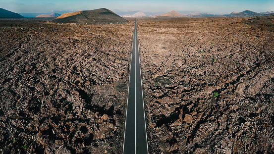 Endless straight highway towards the horizon through Volcanic Landscape. Shot with sunset light. Volcanic Mountains and Hills on the Horizon. Drone Point of View Stitched High Res Panorama DJI Mavic 3 Pro. Timanfaya National Park, Lanzarote Island, Canary Islands, Spain - Africa.