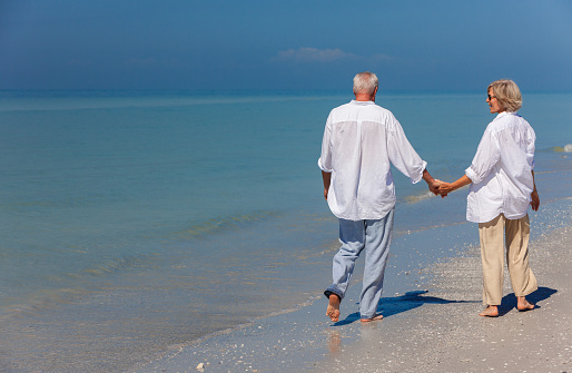 Happy senior man and woman couple walking and holding hands on a deserted tropical beach with bright blue sky