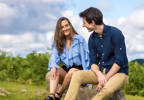Horizontal side view photo of a young elegant couple looking each other sitting on the mountain