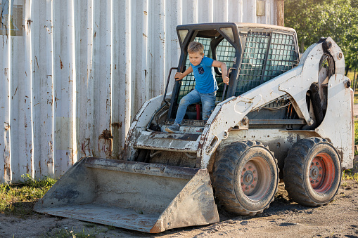 A little old bobcat backhoe in the yard by the garage and a little boy playing in it.