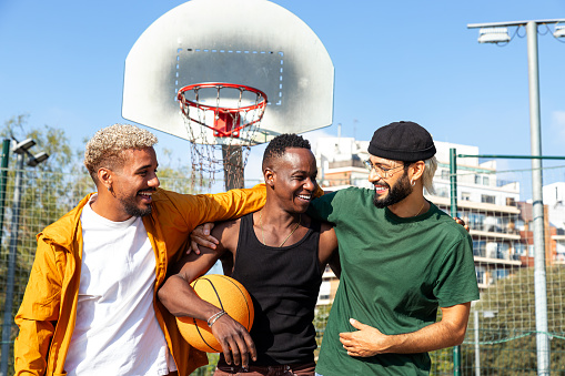 Three happy young multiracial male friends embracing laughing and having fun standing in basketball court outdoors. Sports and friendship concept.