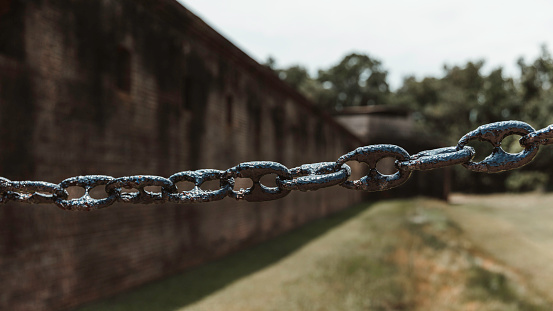 Fort Gaines, Dauphin Island, AL. This evocative photograph showcases a rust-worn chain stretching across the foreground with the historic walls of Fort Gaines in the depth of field. The aged bricks of the fort, standing since the Civil War era, provide a silent testimony to the past. The chain, once bright and functional, now bears the patina of time, symbolizing the strength and endurance of the structures that have witnessed centuries of history.