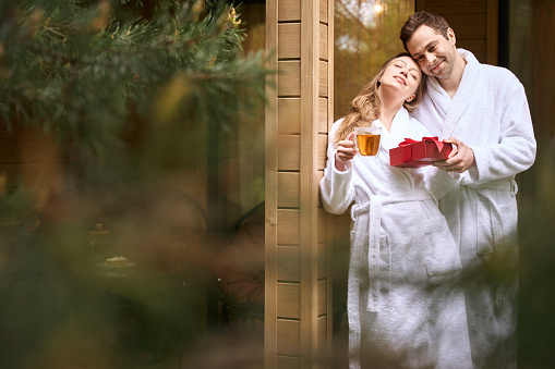 Pretty woman in white bathrobe drinking tea and receiving red box gift from her sweet man, standing on porch