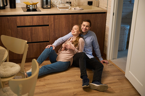 Smiling husband hugging his beautiful pregnant wife while sitting on floor of cozy kitchen