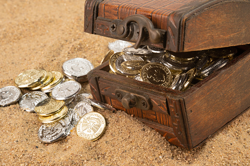 A wooden chest with an ajar lid filled with coins. Coins scattered on the sand.
