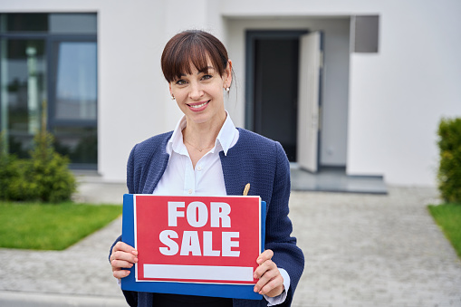 Young woman stands near a country house with a for sale sign, a real estate agent in a business suit