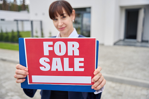Female real estate agent stands with a for sale sign, a lady in a business suit