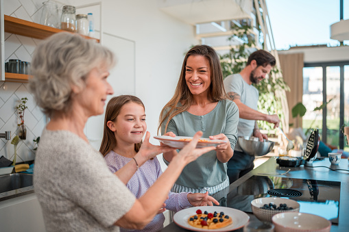 Making waffles in their kitchen, a multigeneration Caucasian family adds a finishing touch with blueberries and raspberries.