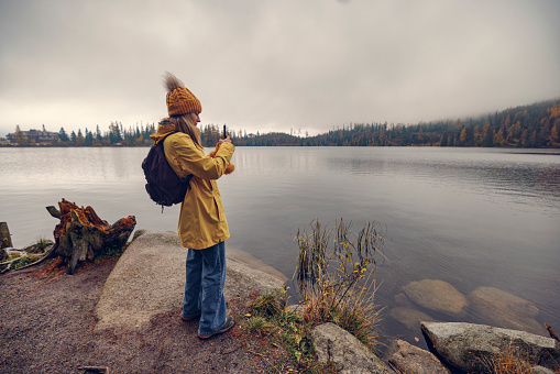 Teenager girl standing on the shore of the lake (Štrbské Pleso) on an autumn day. The girl is filming with her smartphone.\nCanon R5