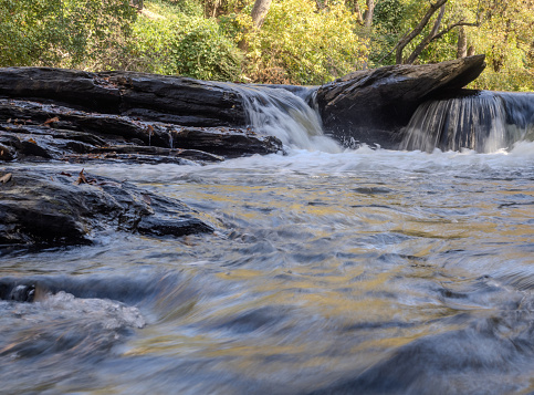 Close up, low angle view of a small waterfall tumbling over plate-like boulders in Big or Vickery Creek in Roswell, Georgia. Deciduous trees are in the background.