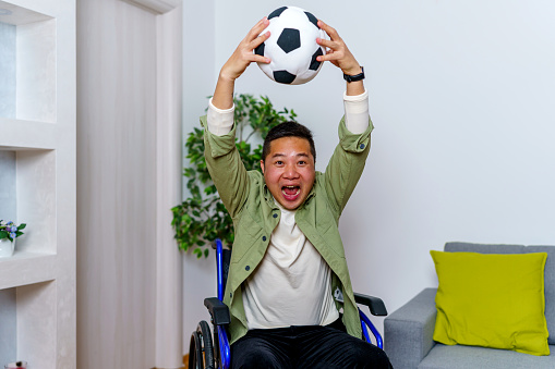 A mid-adult Chinese man in a wheelchair is shown in his living room, engrossed in a soccer game on television, with a soccer ball in hand, a testament to his undiminished love for the sport