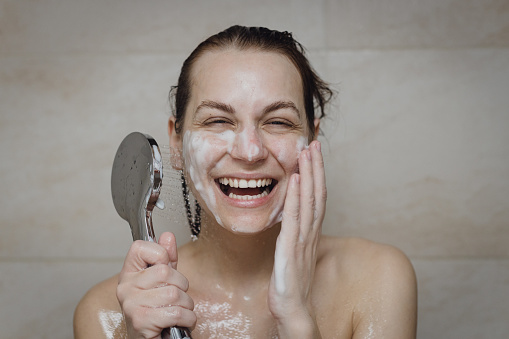 Portrait of a beautiful young woman washing her face with a soap foam and holding a shower head in the bathroom at home