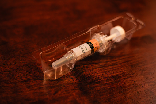 A close-up shot of a prefilled hypodermic syringe ready for use. It is packaged and sealed by the manufacturing pharmaceutical company. In view is a prescribed injection to prevent post-surgical blood clots. The packaged prefilled syringe has been placed on a wooden table. Focus on the barrel. Horizontal format. No people. Copy space. Note to Inspector: Logos and company name have been removed.