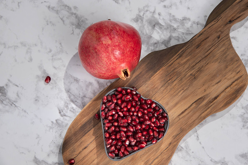 Fresh pomegranate arils in a heart shaped bowl on a wooden cutting board.