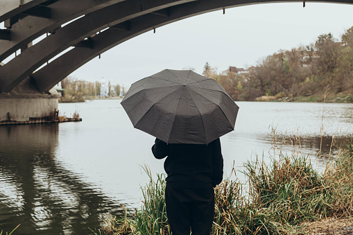 A man with a black umbrella stands by the river in rainy weather, viewed from the back.