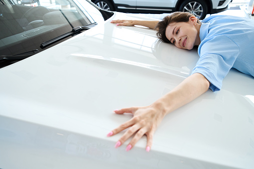 Pretty woman with beautiful manicure hugging hood of auto in car dealership