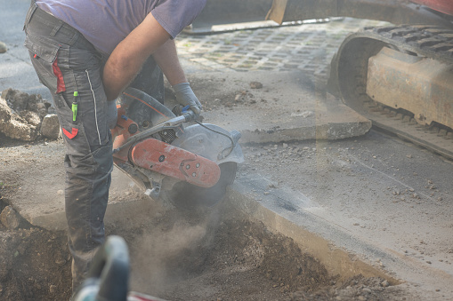 Worker using a motorized asphalt cutter or saw with round blade to cut through the asphalt ground as a process of renovation or remedy of a problem.