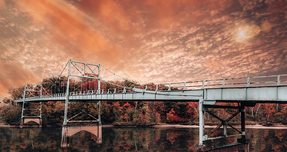 The image presents a dramatic and fiery sky casting its vibrant hues over an old suspension bridge. The rich colors of the sunset reflect on the calm waters below, creating a mirror-like effect that doubles the intensity of the scene. The bridge, a symbol of connection and passage, stands out against the backdrop of changing autumn leaves, offering a nostalgic blend of human engineering and natural splendor.