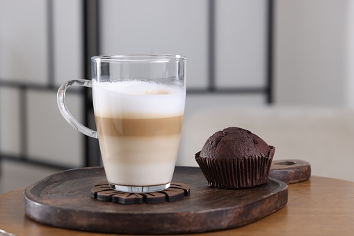 Cup of aromatic latte macchiato and chocolate muffin on wooden table against blurred background