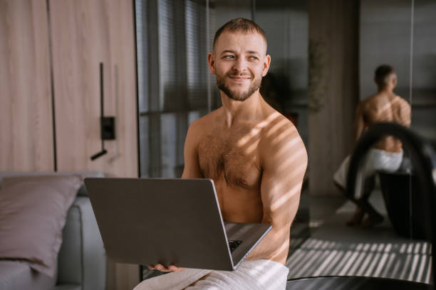 istock Young bearded caucasian man with wrapped towel around hips sitting at hotel using laptop at morning smile looks at camera. Confident European guy at home remote working. Home office. 1815401246
