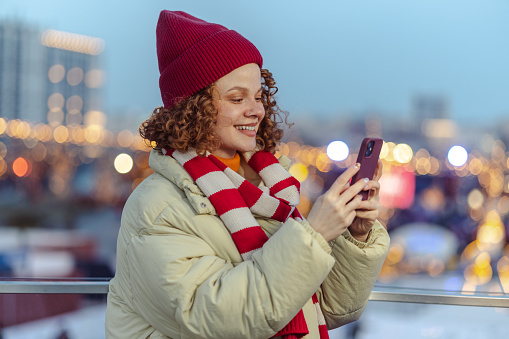 Smiling redhead woman wearing warm clothing and enjoying Christmas time in the city. She is using smart phone and smiling