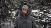 A young woman stands against a background of fir trees under heavy snowfall in winter. A girl in glasses and a down jacket with a hood stands and looks at the camera. Closeup.