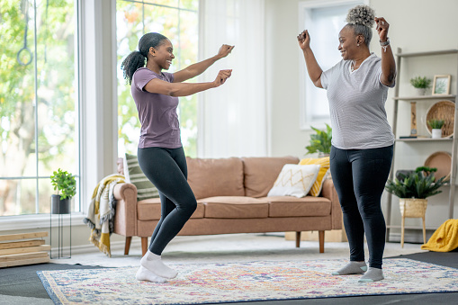 A Mother and her adult daughter are. seen dancing in the living room together as they spend time together.  They are both dressed comfortably and are laughing as they have fun connecting with one another.