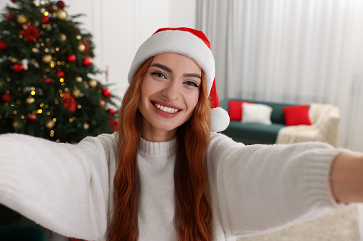 Beautiful young woman in Santa hat taking selfie at home. Christmas celebration