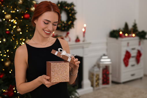 Happy young woman opening gift box in room decorated for Christmas, space for text