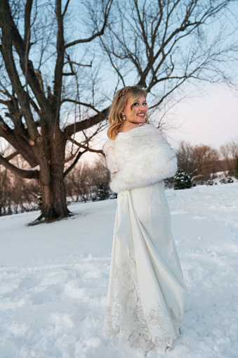 Backview of a blond bride in white wedding dress and fur stole walking  across snow in snowshoes, Minnesota