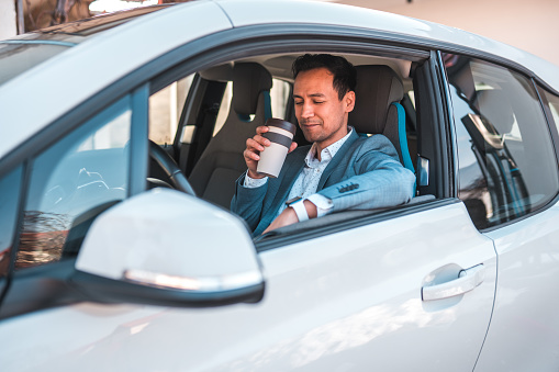 Asian man enjoys his beverage while seated in the driver's seat of a modern electric car. He is dresses professionally on a suit.