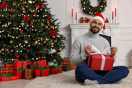 Happy young man in Santa hat opening Christmas gift at home