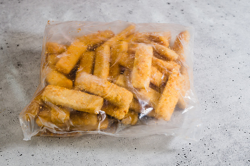 Bag of frozen breaded fish sticks close-up on grey stone background