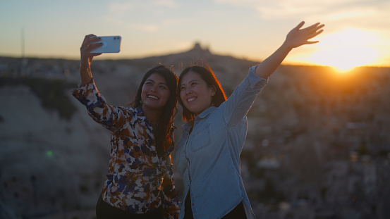 Two female tourist friends are using a mobile smart phone to take selfies and capture memories during the sunset from their travel in Cappadocia Türkiye Turkey