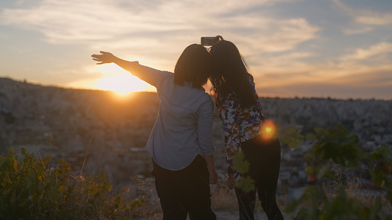 Two female tourist friends are using a mobile smart phone to take selfies and capture memories during the sunset from their travel in Cappadocia Türkiye Turkey