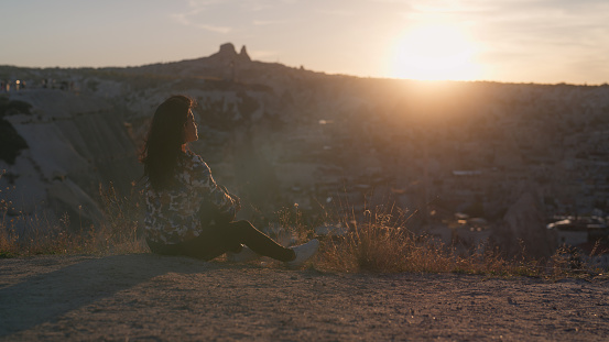 A multiracial female tourist is sitting on ground relaxing and watching the sunset on top of hill in Cappadocia in Türkiye Turkey.