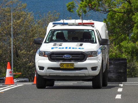 A paddy wagon of the New South Wales Police parked on Bondi Road near Bondi Beach. The Police closed off Bondi Road after a traffic accident involving several cars and a scooter.  The traffic cones have the logo of the NSW Government. In the distance is the Pacific Ocean.  At the bottom of this hill and to the left is Bondi Beach.  This image was taken on the corner of Dudley Street and Bondi Road on a sunny afternoon on 26 November 2023.