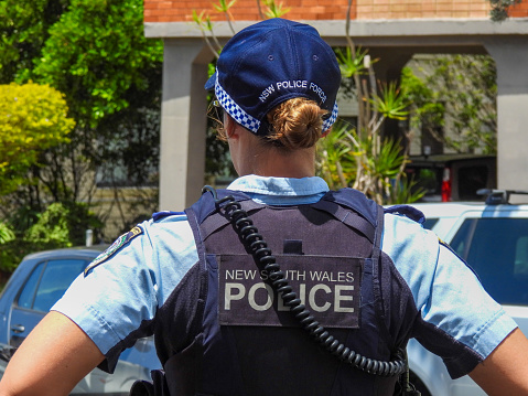 A Senior Constable of the New South Wales Police in attendance at the site of a traffic accident involving several cars and a scooter.  She is looking in the direction of the damaged cars and scooter.  This image was taken near the corner of Dudley Street and Bondi Road on a sunny afternoon on 26 November 2023.