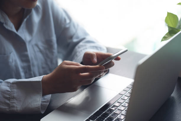 istock Business woman sitting at office table using mobile phone while online working on laptop computer at home office, business casual, telecommuting, working from home, close up 1815793322